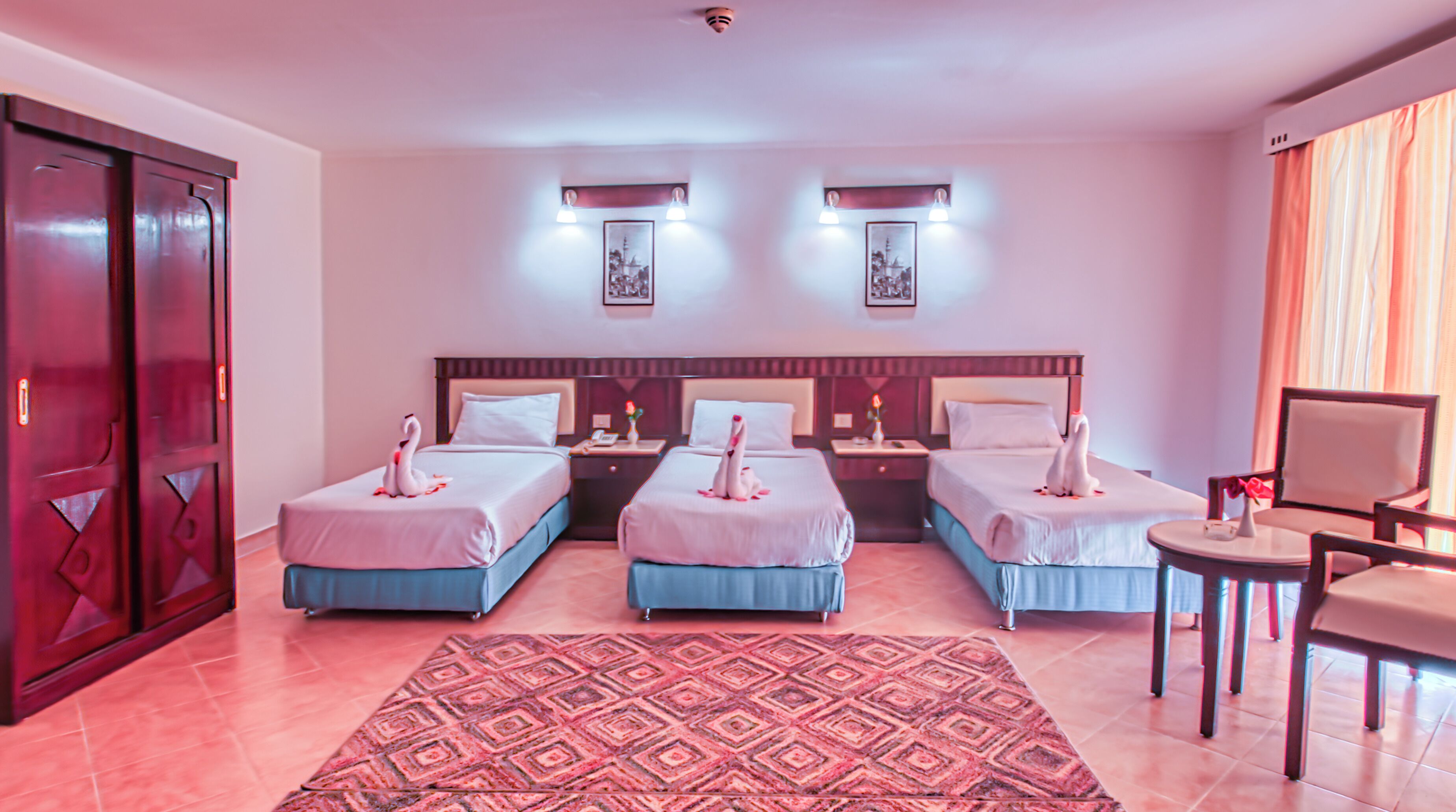 A hotel room with three beds, a desk, and a chair. There are two wall-mounted lamps and a large patterned rug.