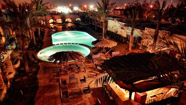 Vista nocturna de un resort con dos piscinas, sillas de descanso, palmeras y una zona de bar.