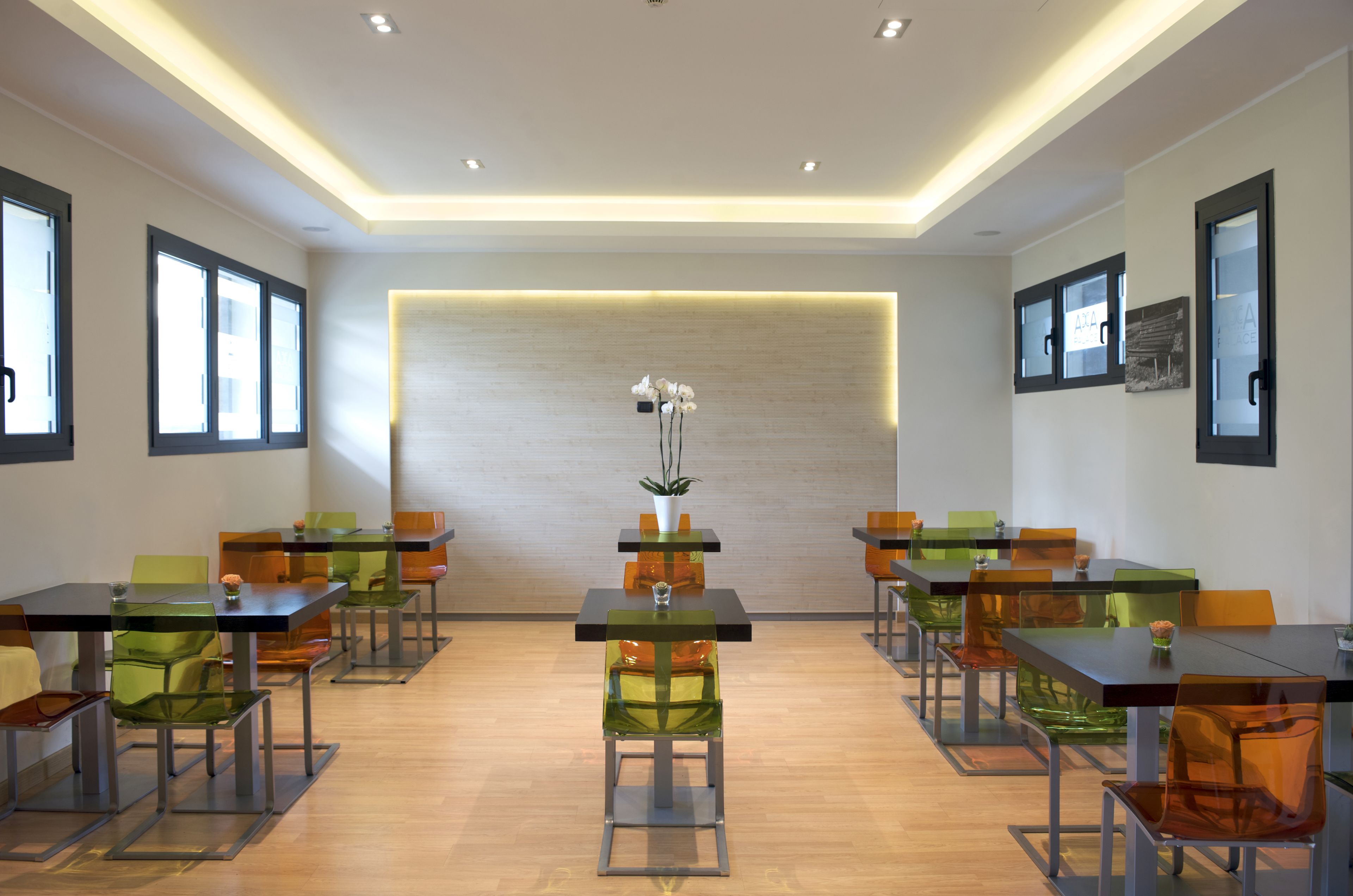 A modern dining area with multiple tables and chairs, a central decorative plant, and a neutral-toned wall with recessed lighting.