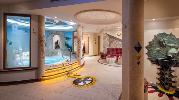 A luxurious indoor pool area with a glass wall, a hot tub, and a decorative sculpture. - Hotel Alpenjuwel Jäger (Tux)