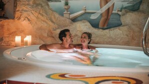 A couple in a hot tub with a mural of a woman in a swimsuit. There are candles on the side. - Hotel Alpenjuwel Jäger (Tux)