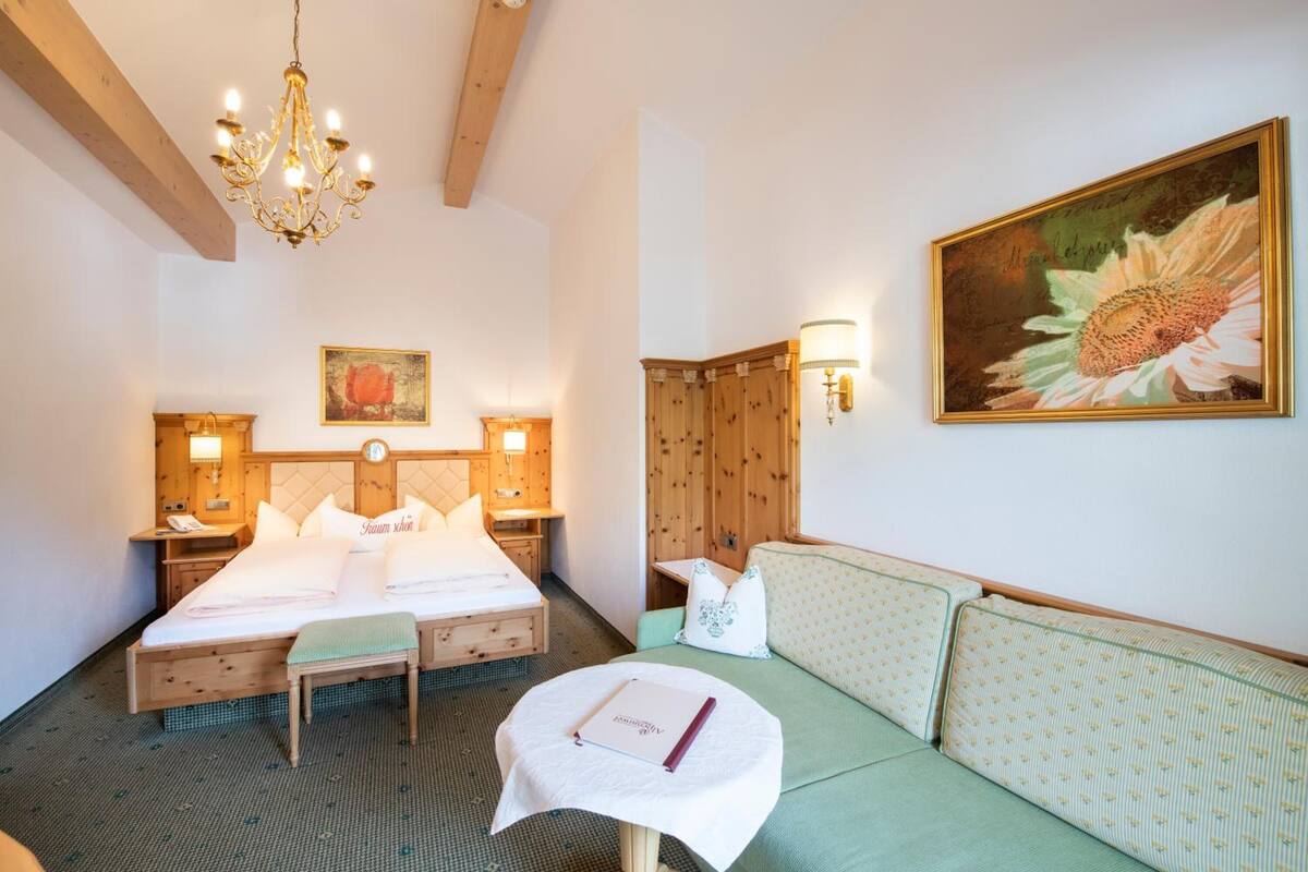 Ein Hotelzimmer mit einem Bett, einem grünen Sofa, einem kleinen Tisch mit einem Buch, einer Kandelaber, einem Gemälde und Holzvertäfelung.