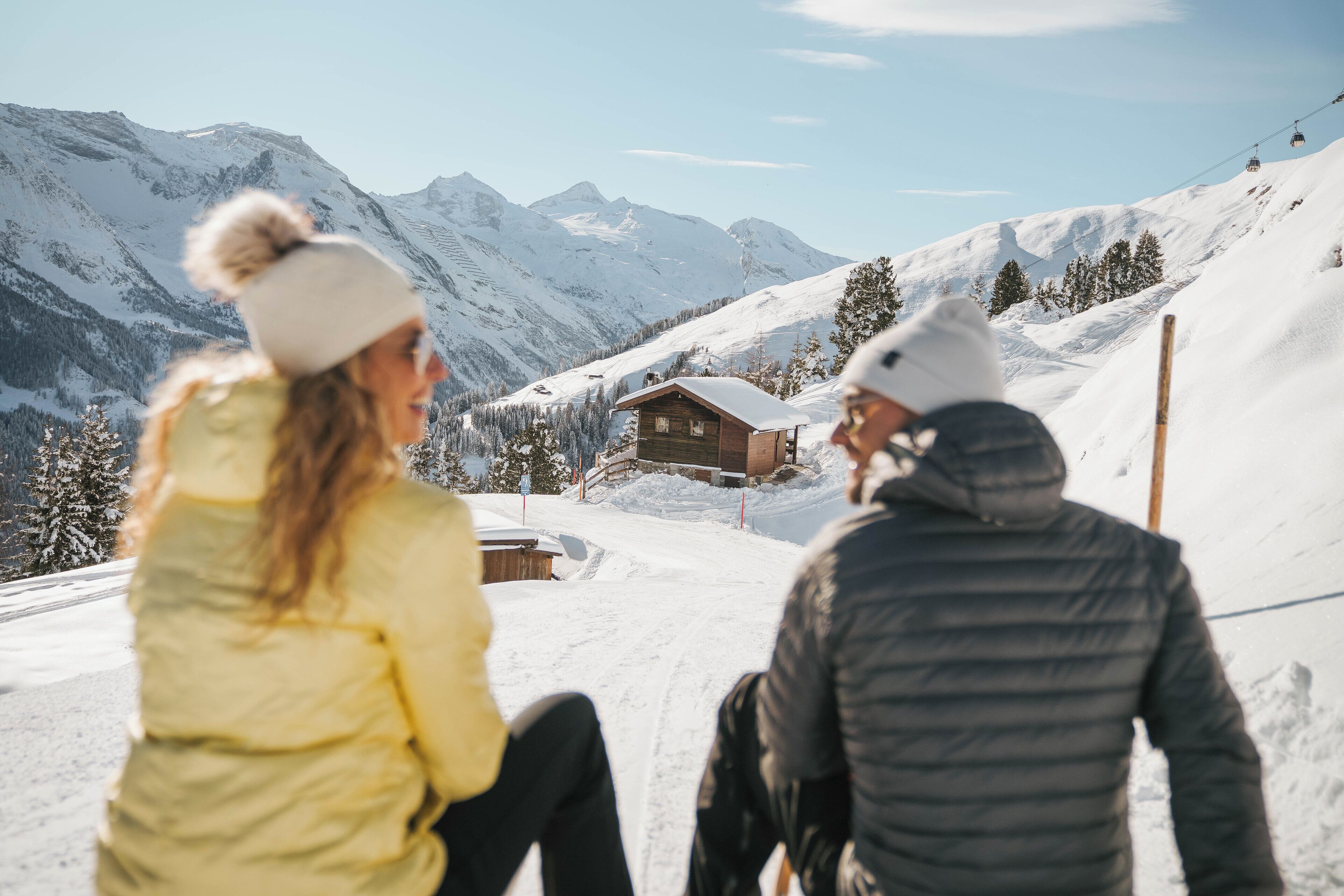 Two people sitting on a snow-covered slope with a wooden cabin and mountains in the background.