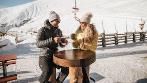Two people toasting at a wooden table with a snowy mountain backdrop. - Hotel Alpenjuwel Jäger (Tux)