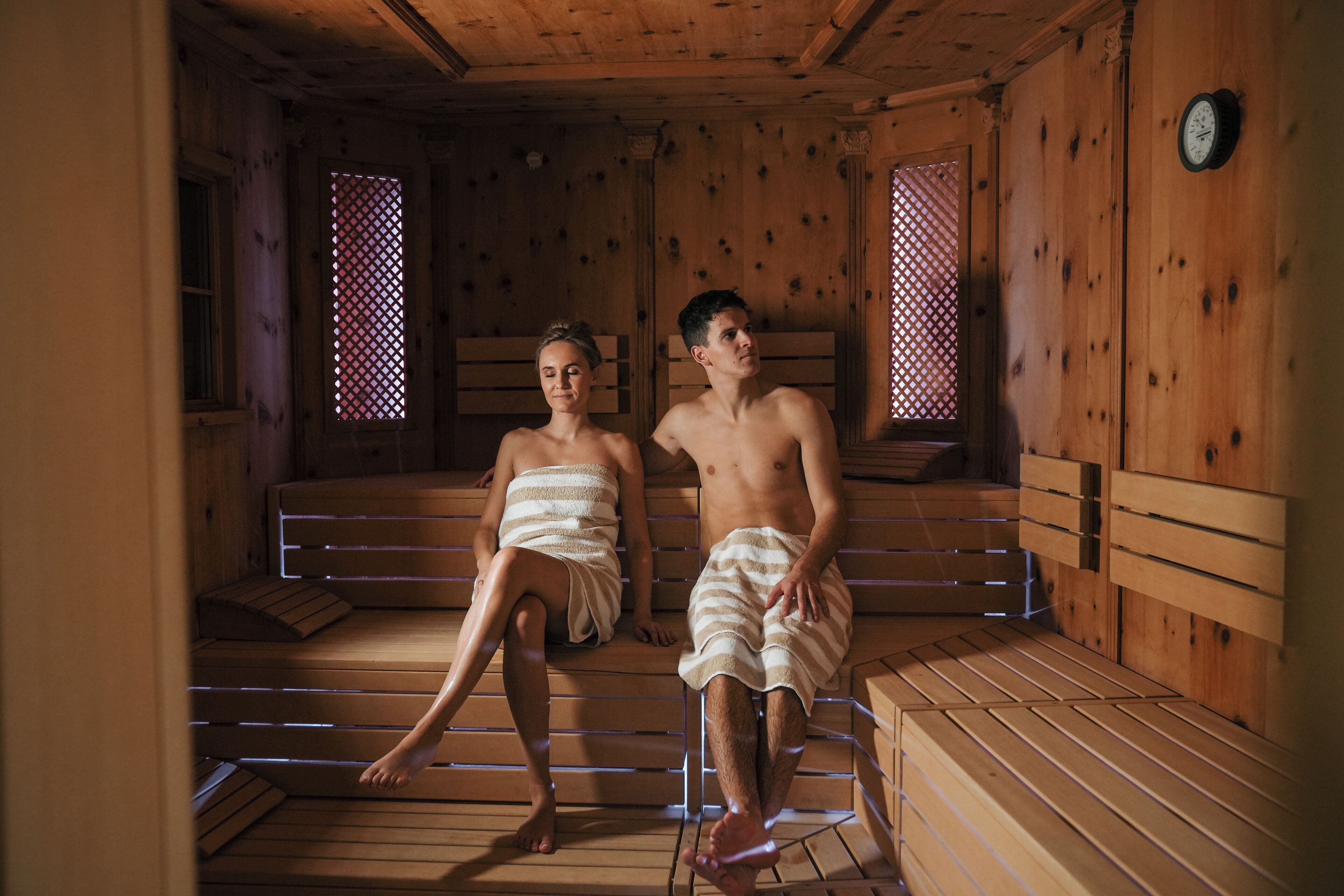 Dos personas en una sauna con bancos de madera y ventanas de celosía.