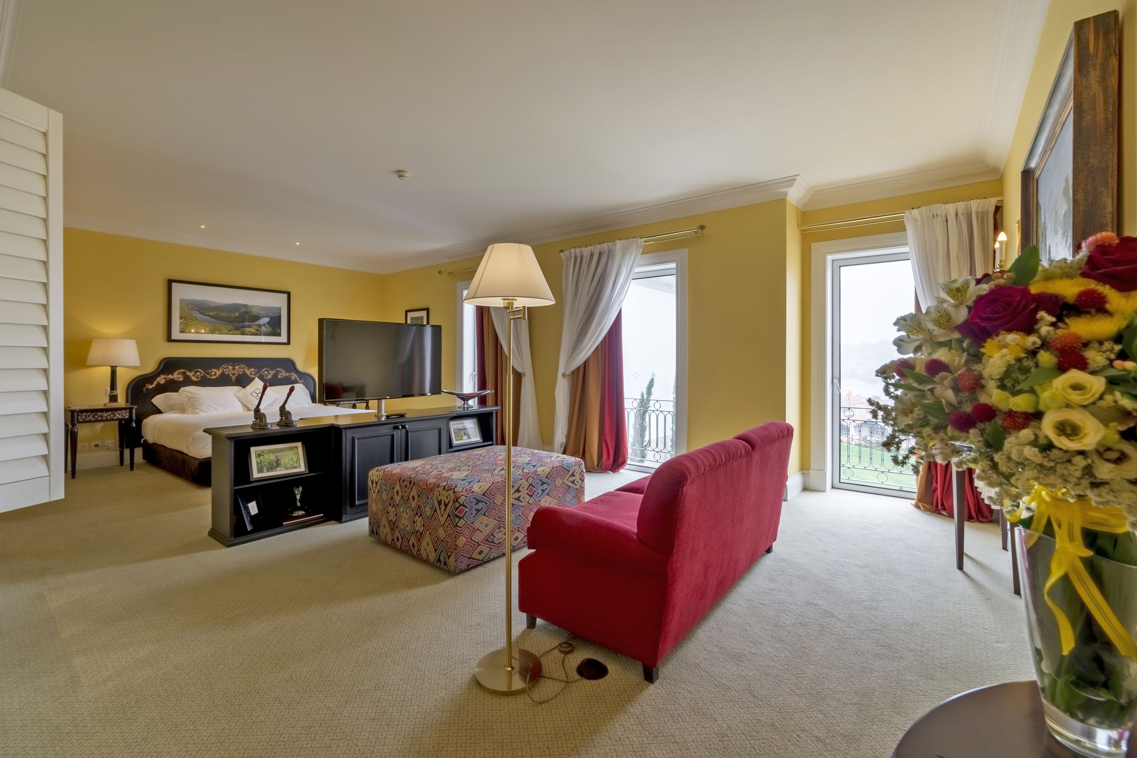 A hotel room with a bed, a desk, a red armchair, a floor lamp, and a large window with curtains.