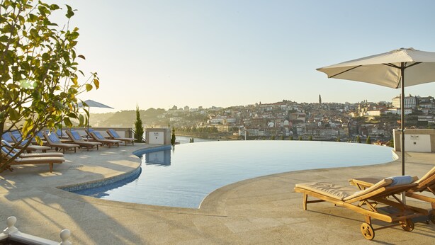 Piscina infinita en la azotea con sillas de descanso y sombrillas, con vista a un paisaje urbano.