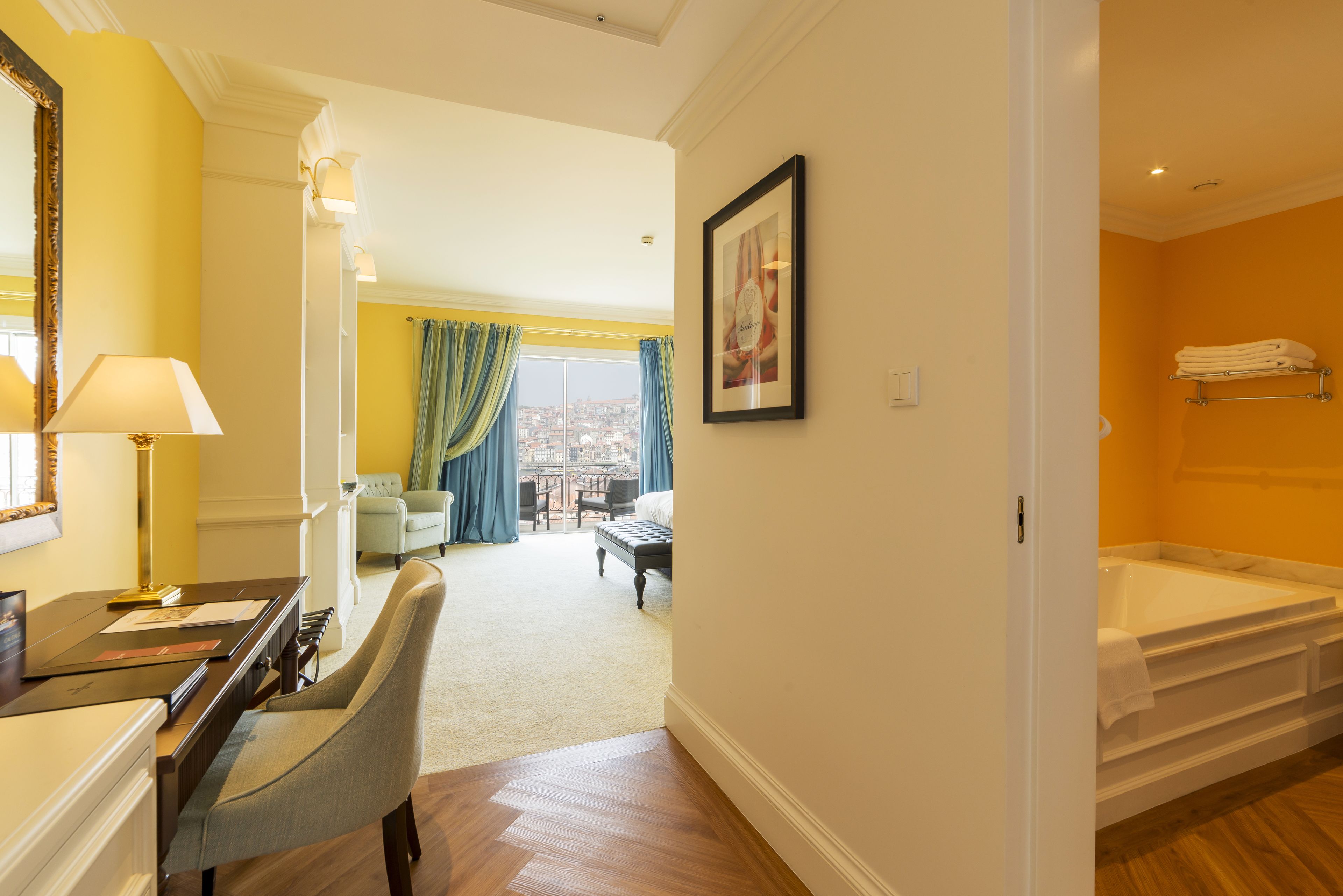 A hotel room with a wooden floor, a desk with a lamp, a chair, a bed, a bathtub, and a view of the city through large windows.
