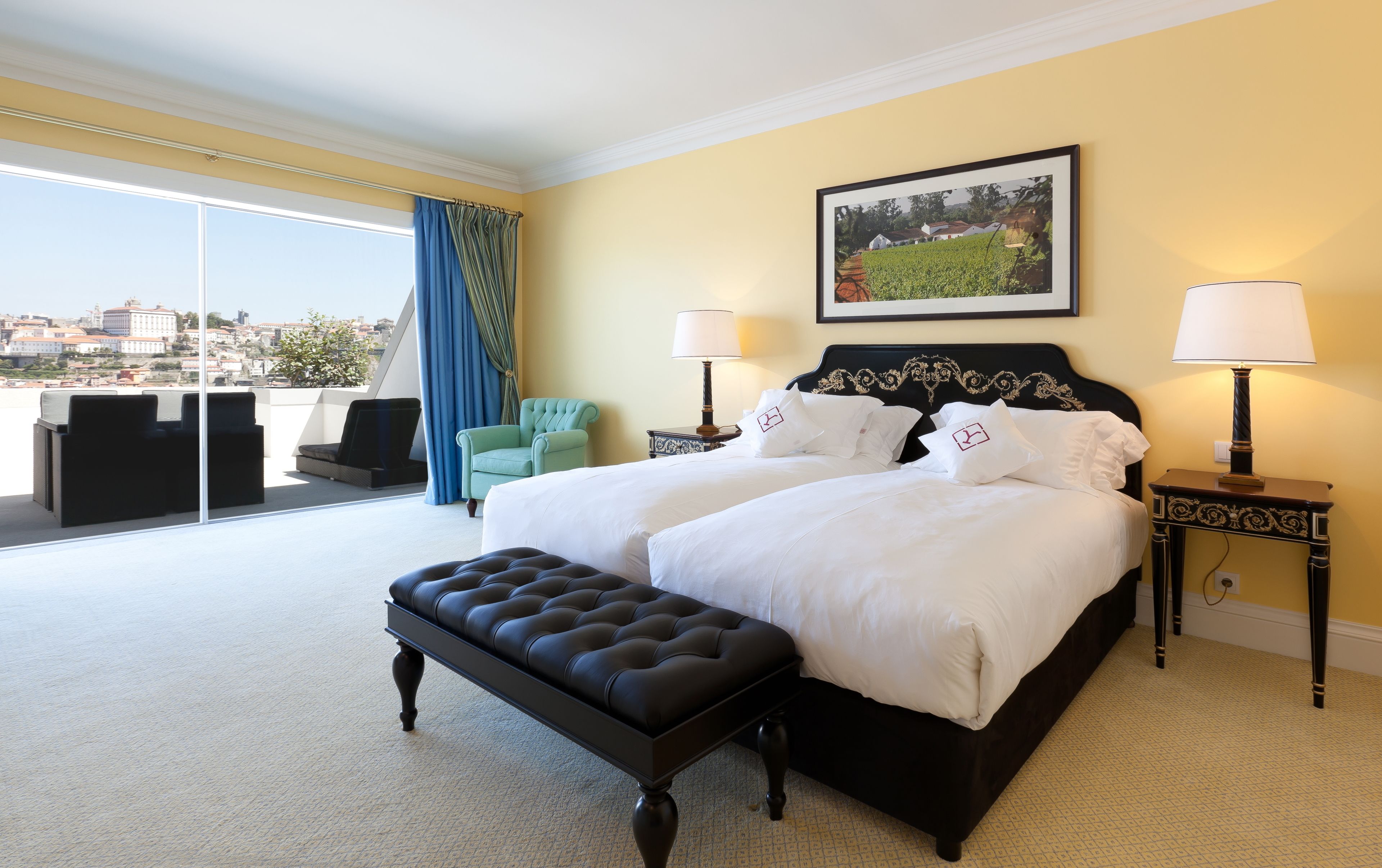 A hotel room with a large bed, two bedside tables with lamps, a green armchair, a black ottoman, and a city view through floor-to-ceiling windows.