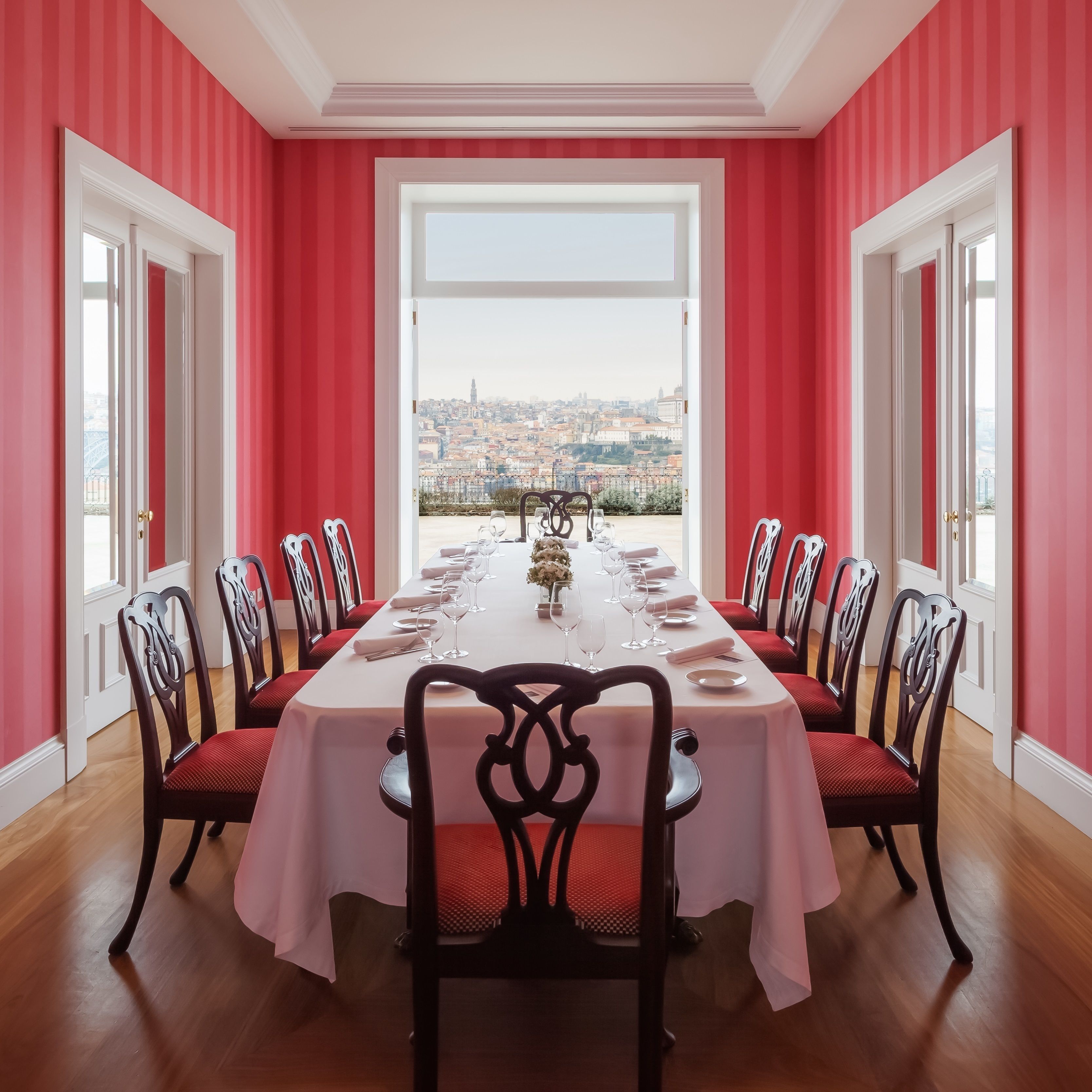 A dining room with a long table set for a meal, flanked by chairs, with a city view through a large window.
