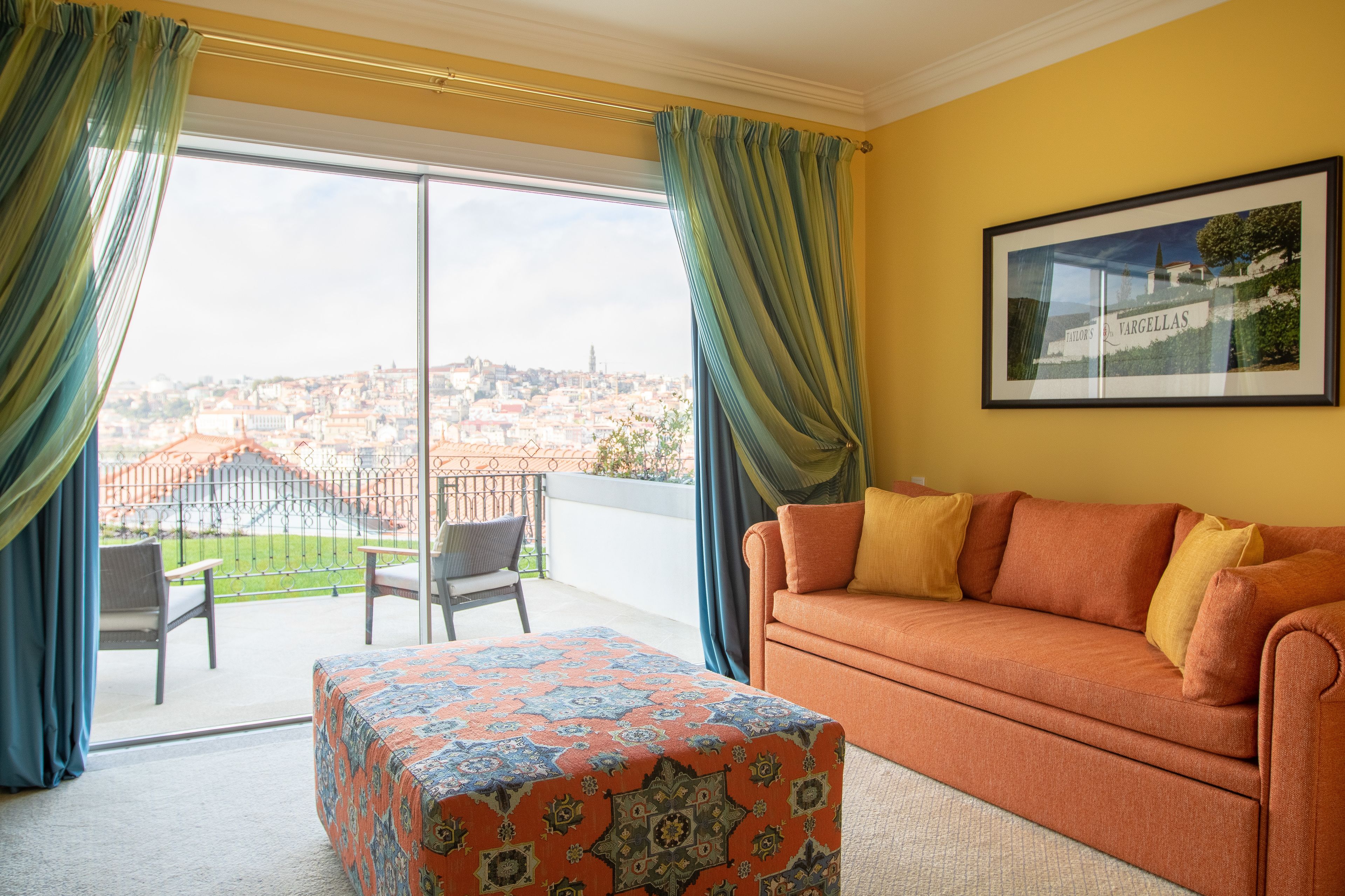 A room with a sofa, a patterned ottoman, and a large window with curtains.