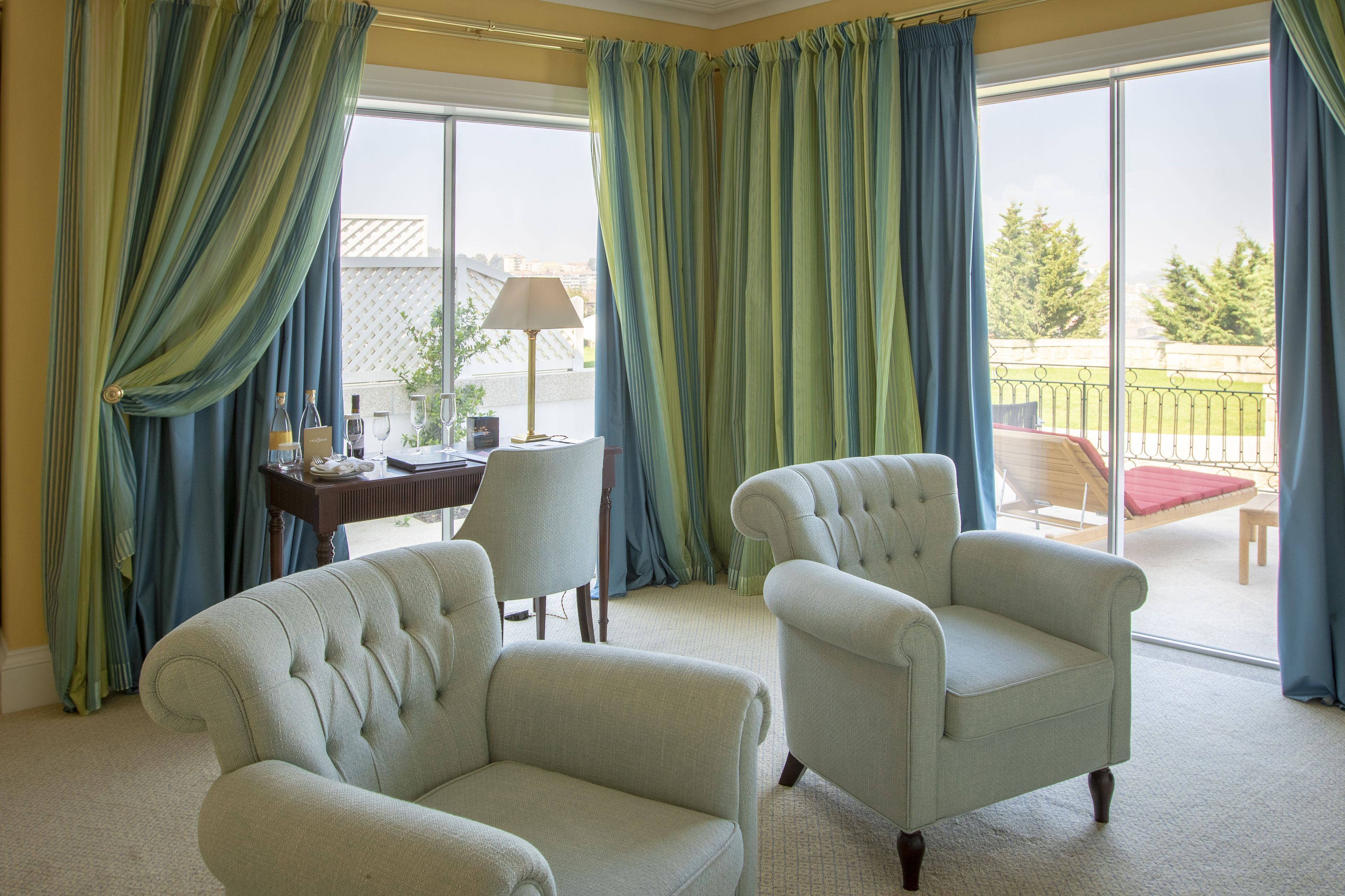 A room with two upholstered armchairs, a table with a lamp, and a view of a balcony with outdoor furniture.