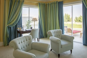 A room with two upholstered armchairs, a table with a lamp, and a view of a balcony with outdoor furniture.