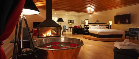 A modern hotel room with a central heated floor, a bed, a seating area, and a fireplace.