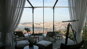 Una habitación soleada con vistas a un paisaje urbano, amueblada con sillas de mimbre y una mesa, y un telescopio sobre un trípode.