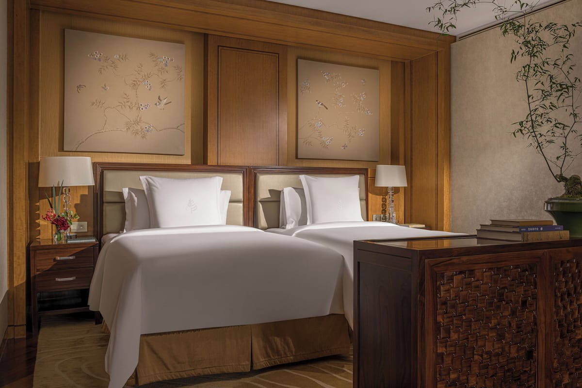 A hotel room with two beds, wooden headboards, a nightstand with a lamp, and a decorative wall panel.