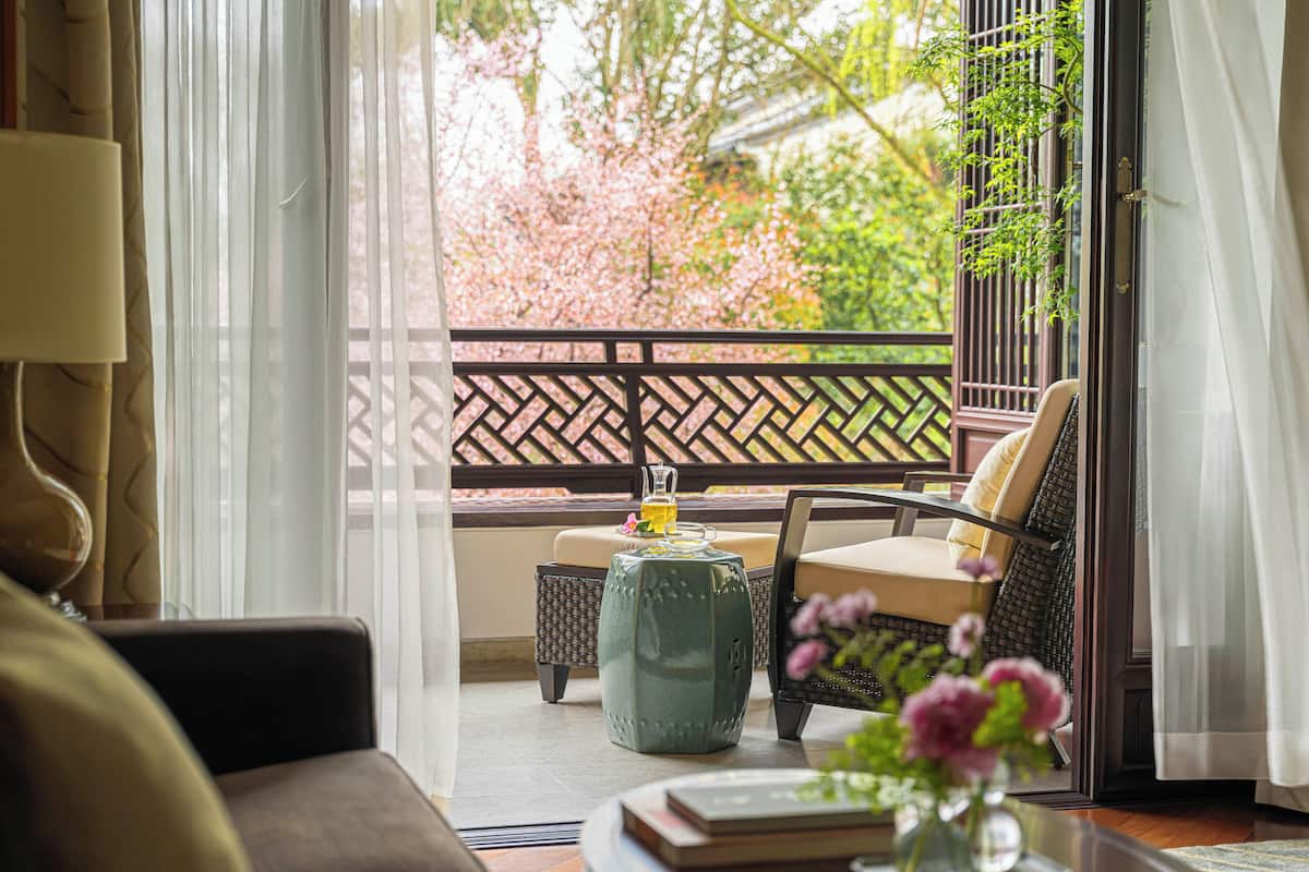 A room with a view of cherry blossoms, a sofa, a chair, a small table with a vase, and a laptop.