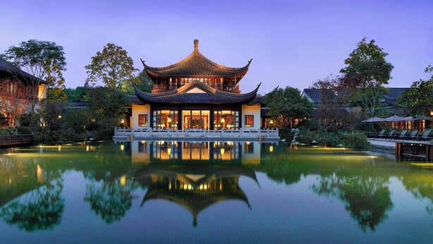 A traditional Chinese building with a reflection in the water, surrounded by trees and a wooden bridge.