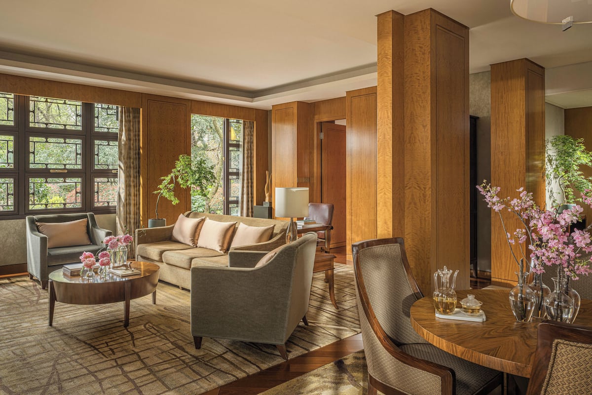 A spacious living room with a sofa, armchairs, a coffee table, and a dining table with chairs. Large windows provide natural light and views of greenery outside.
