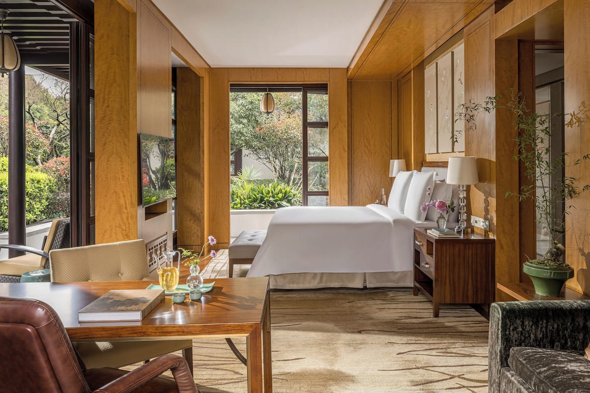 A hotel room with a bed, a desk, a chair, a nightstand, a lamp, and a potted plant.