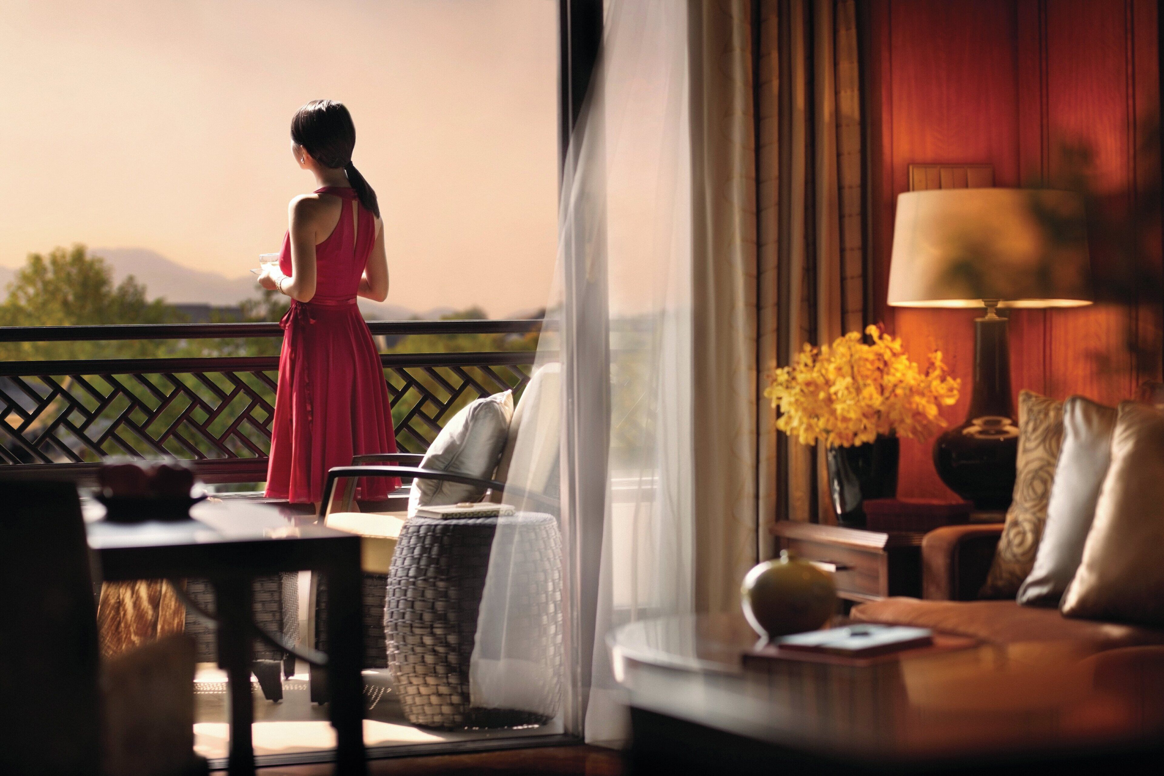 A woman in a red dress standing by a balcony with a view of mountains. Inside, there is a sofa, a lamp, and a vase with flowers.