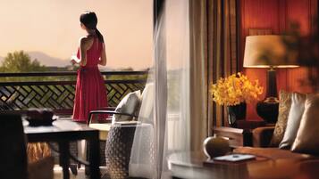 A woman in a red dress standing by a balcony with a view of mountains. Inside, there is a sofa, a lamp, and a vase with flowers.