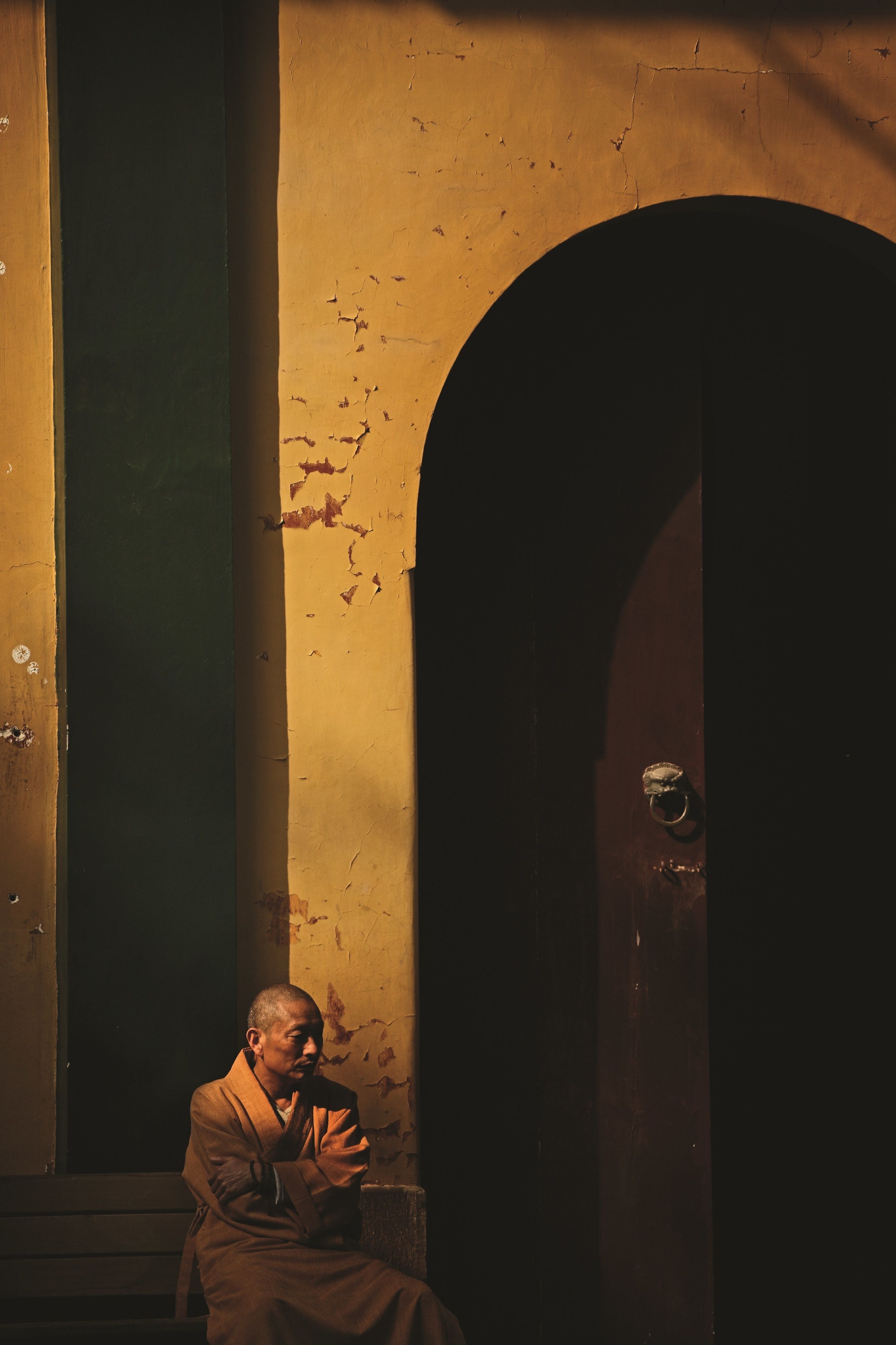 Una persona sentada en un banco en una habitación tenuemente iluminada, con pintura descascarada en las paredes y una puerta de madera.