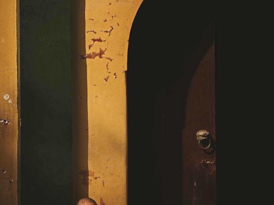 Una persona sentada en un banco en una habitación tenuemente iluminada, con pintura descascarada en las paredes y una puerta de madera.