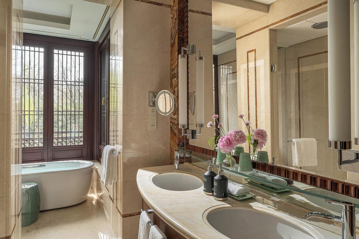 A modern bathroom with a freestanding bathtub, two sinks, a large mirror, and a view through a window with a wooden door.