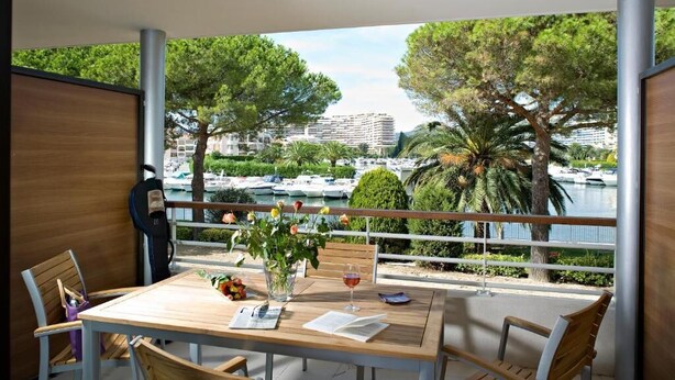 A balcony with a wooden table, chairs, and a vase with flowers. There is a view of a marina with boats and buildings in the background.