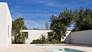A swimming pool with clear blue water, surrounded by white walls and greenery, including palm trees and shrubs.