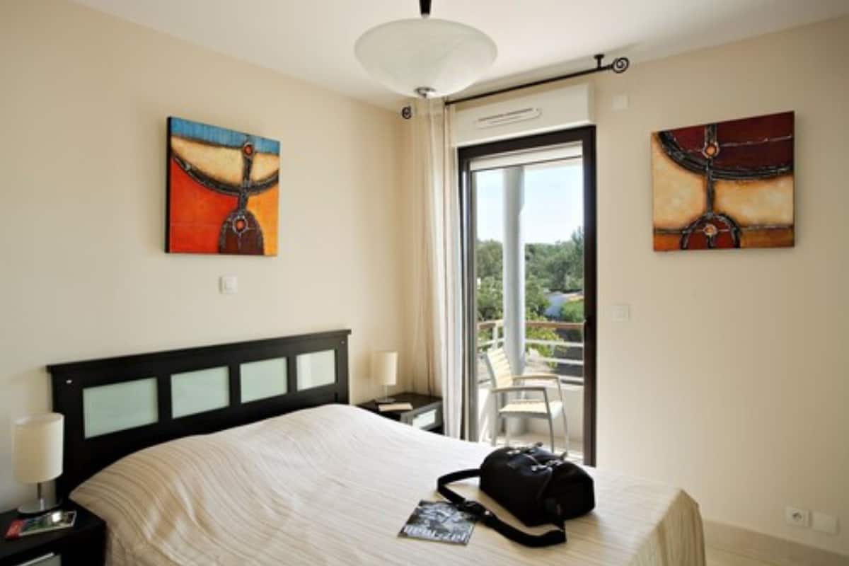 A hotel room with a bed, a nightstand, a lamp, a window with a view, and two paintings on the walls.