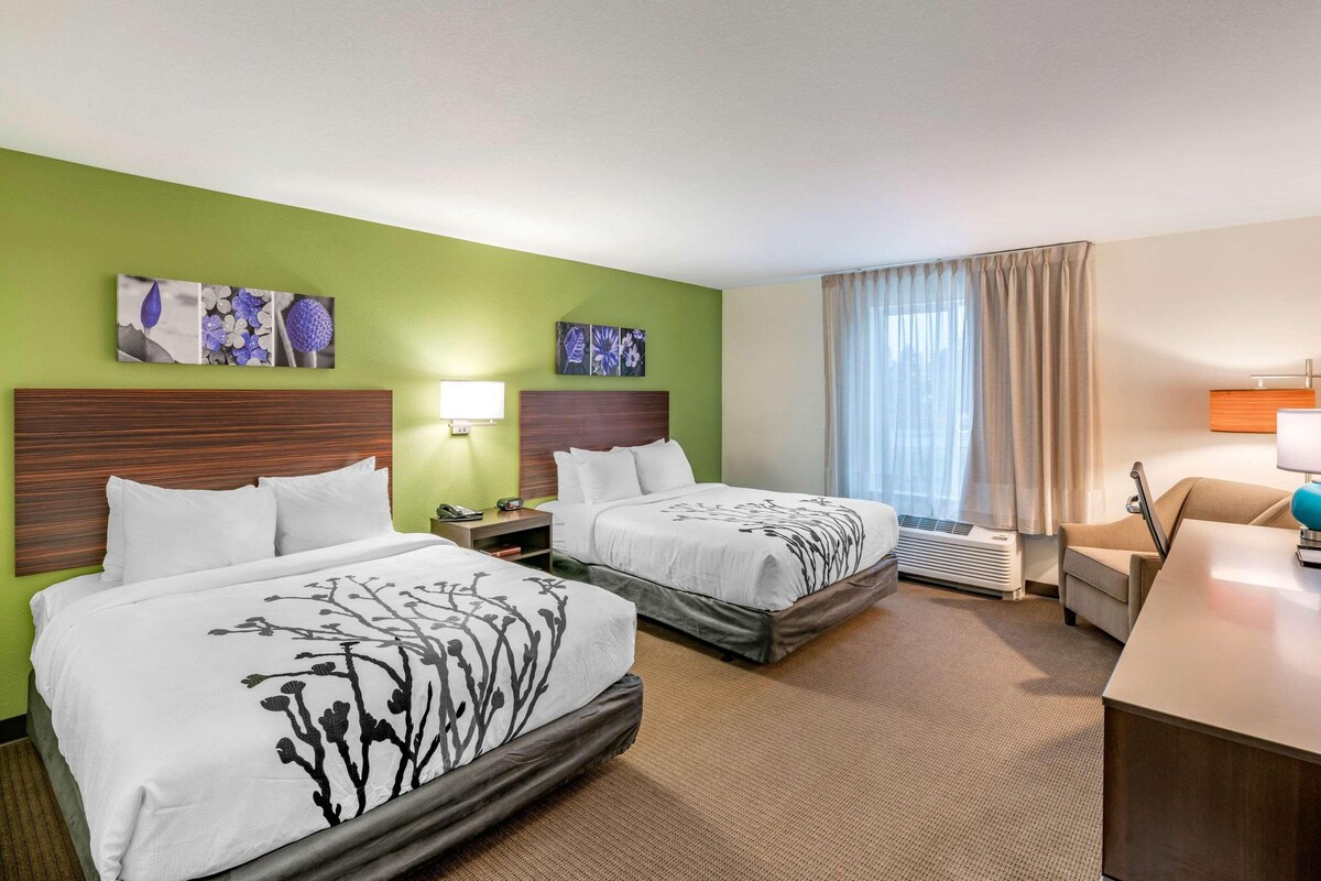 A hotel room with two beds, a desk, a chair, a nightstand, and a window with curtains.