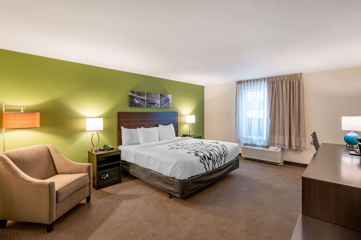 Ein Hotelzimmer mit Bett, Sessel, Schreibtisch, Lampe, Heizung und einem Fenster mit Vorhängen.