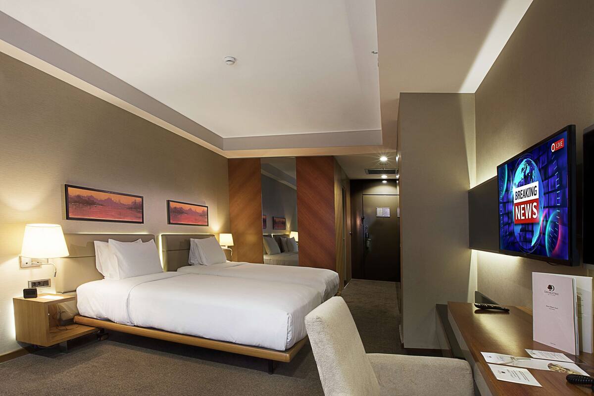 A hotel room with a large bed, two bedside tables with lamps, a flat-screen TV mounted on the wall, and a desk with papers and a remote control.