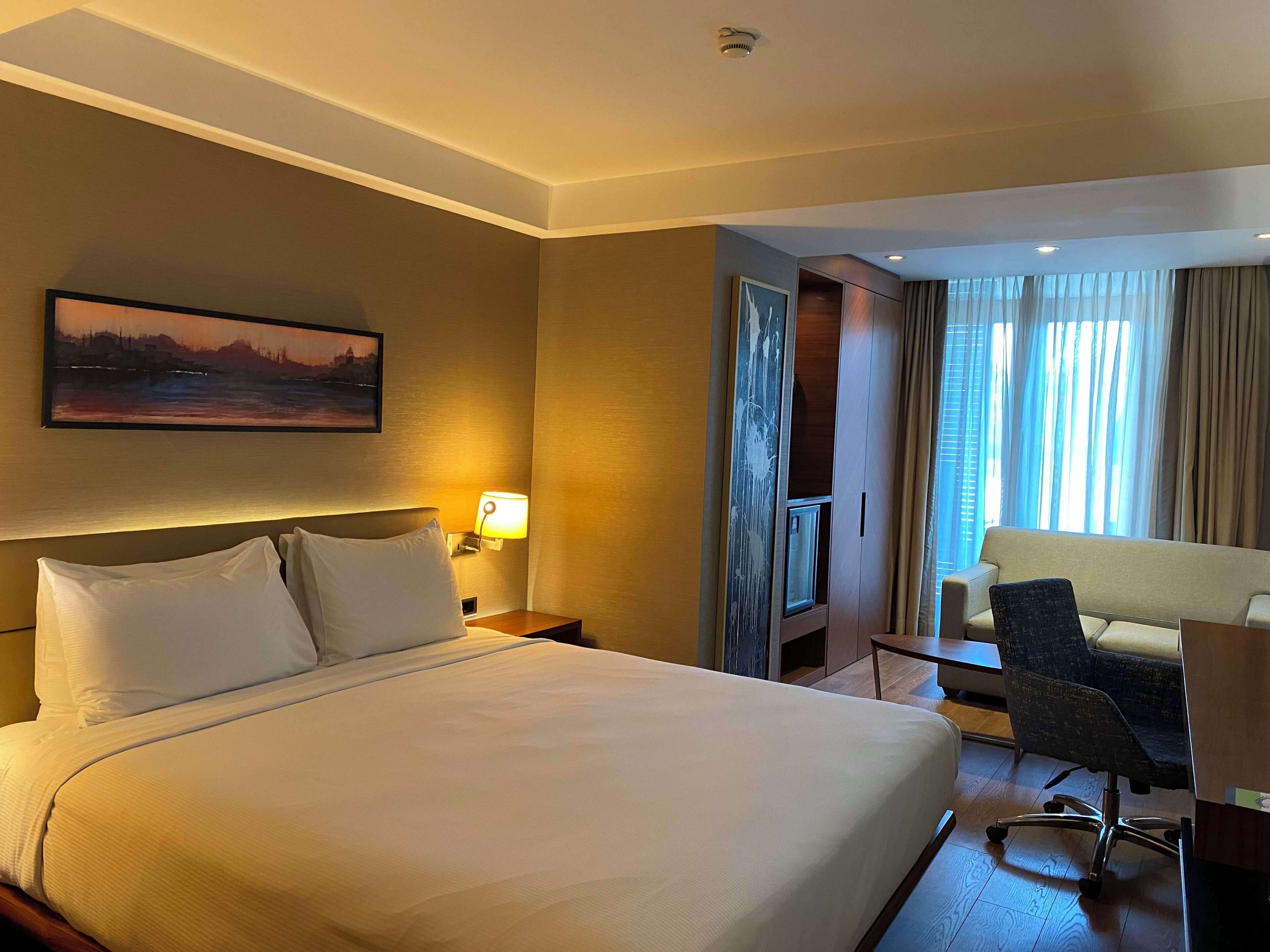 Una habitación de hotel con una cama grande, un escritorio, una silla, un televisor y un cuadro en la pared.