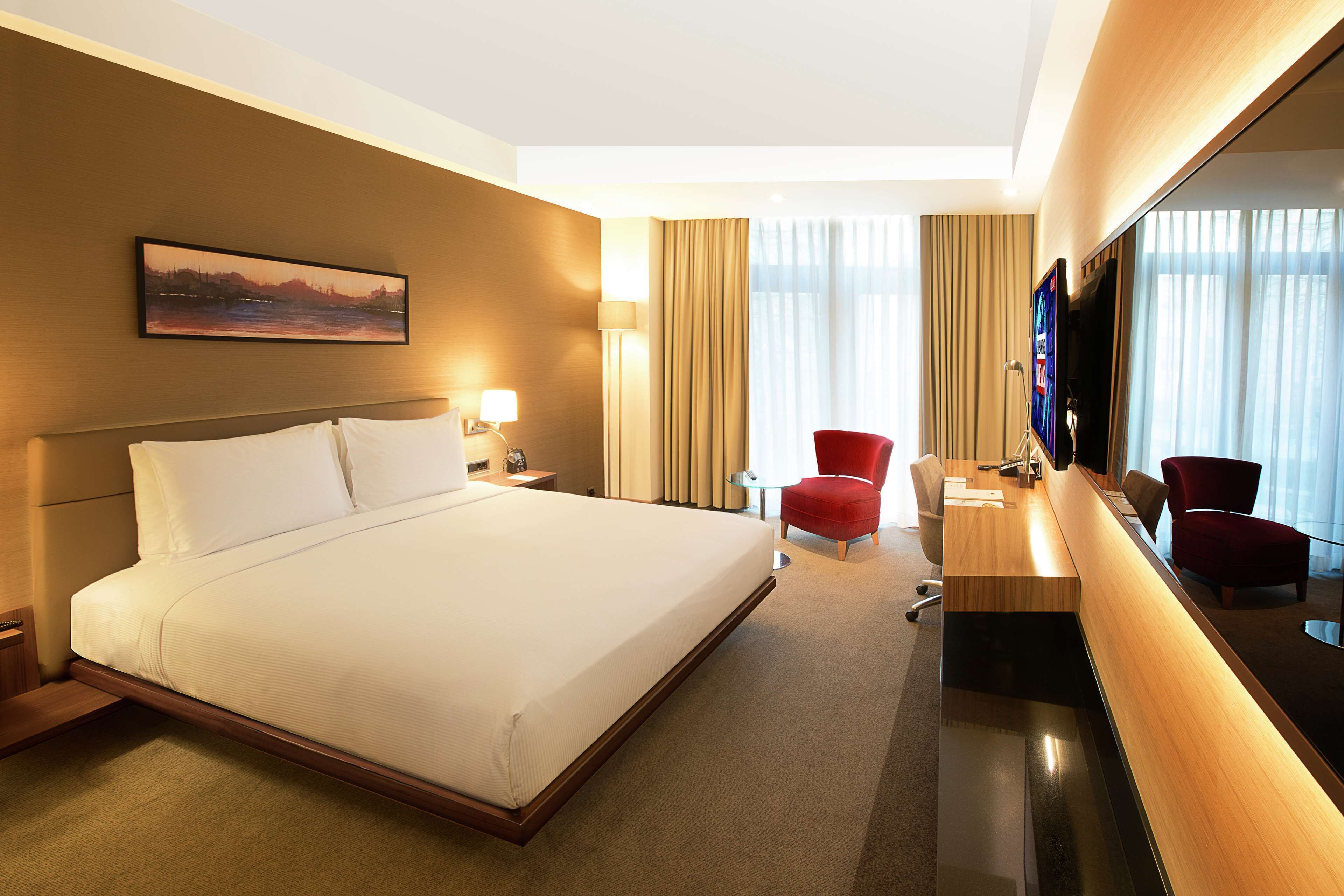 A hotel room with a large bed, a desk, a TV, and a red chair.