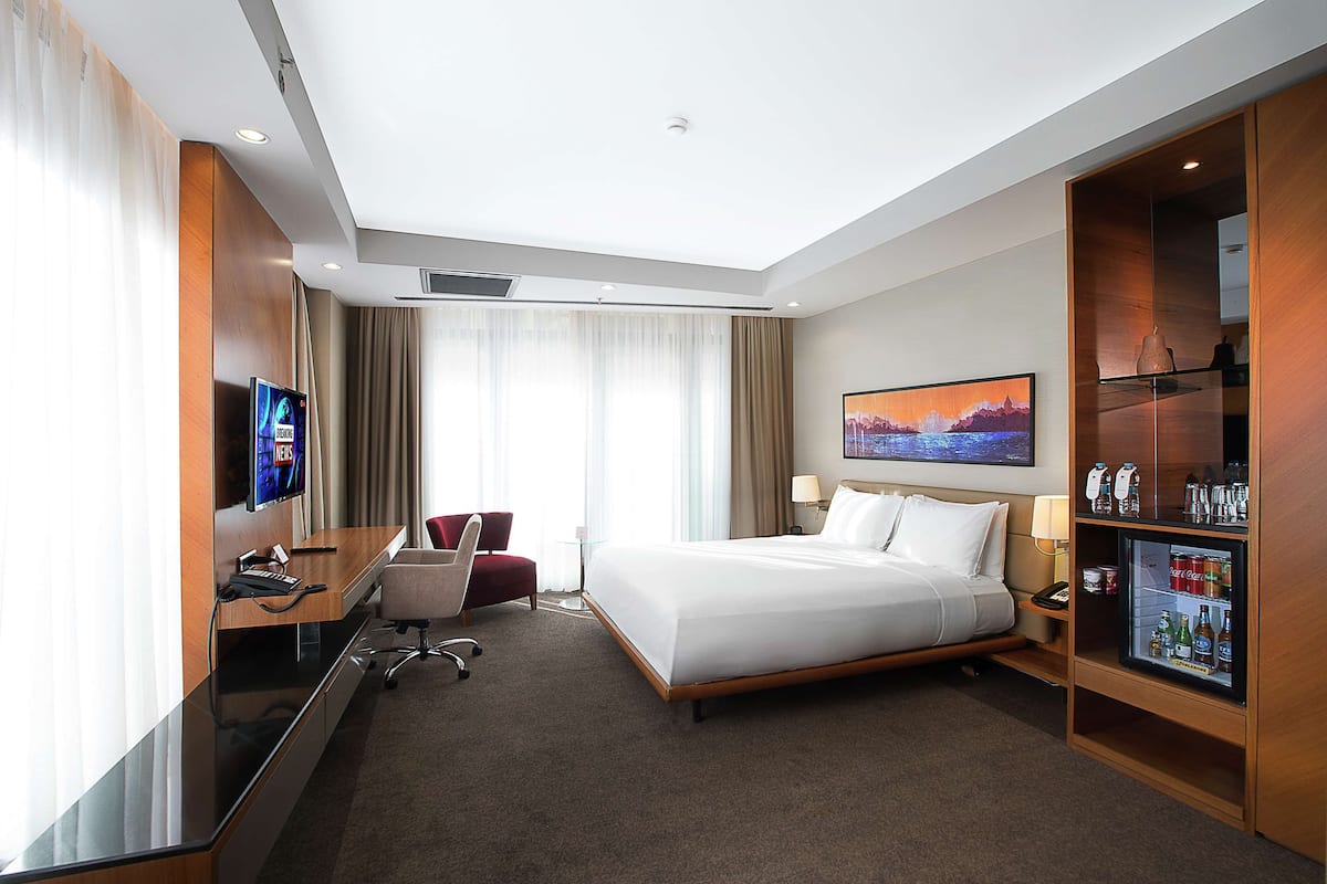 A hotel room with a bed, a desk with a computer, a TV, a refrigerator, and a window with curtains.