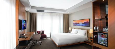 Une chambre d’hôtel équipée d’un lit, d’un bureau, d’une chaise, d’une télévision, d’un mini-bar et d’une grande fenêtre.