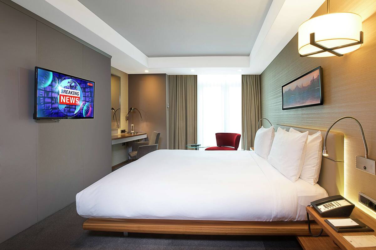 A hotel room with a large bed, a TV displaying news, a desk with a lamp, a chair, and a bedside table with a phone and papers.