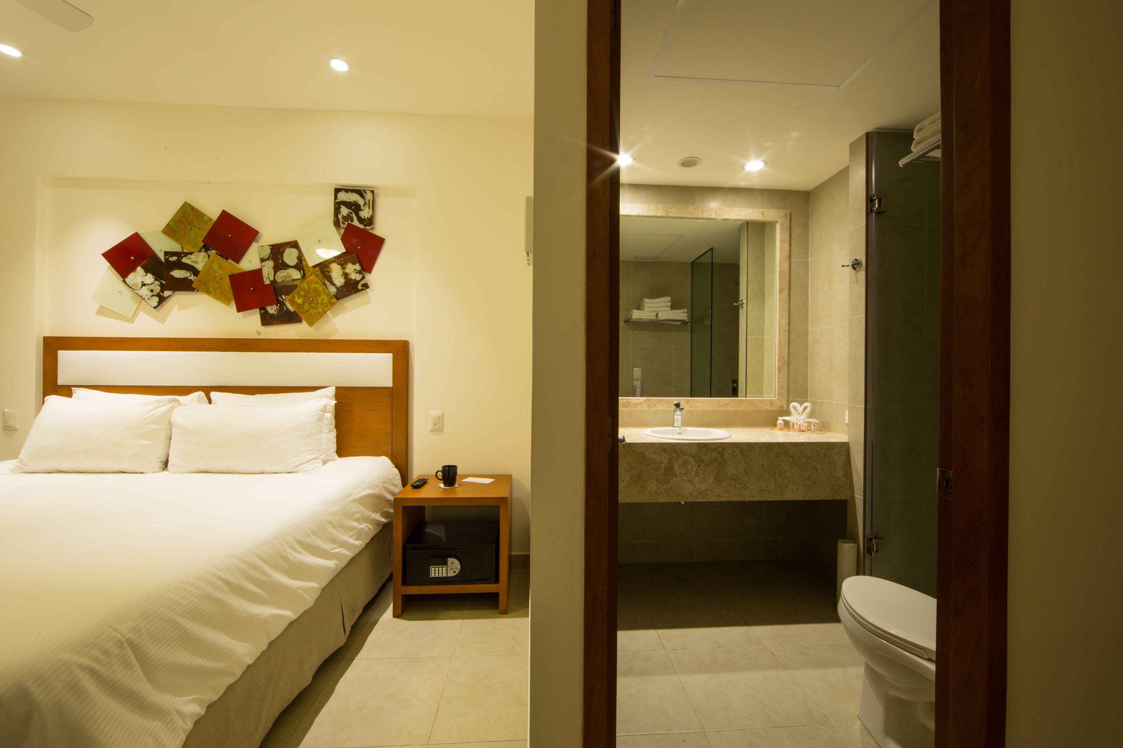 Habitación de hotel con cama, mesita de noche, una obra de arte en la pared, baño con lavamanos y espejo, y un inodoro.