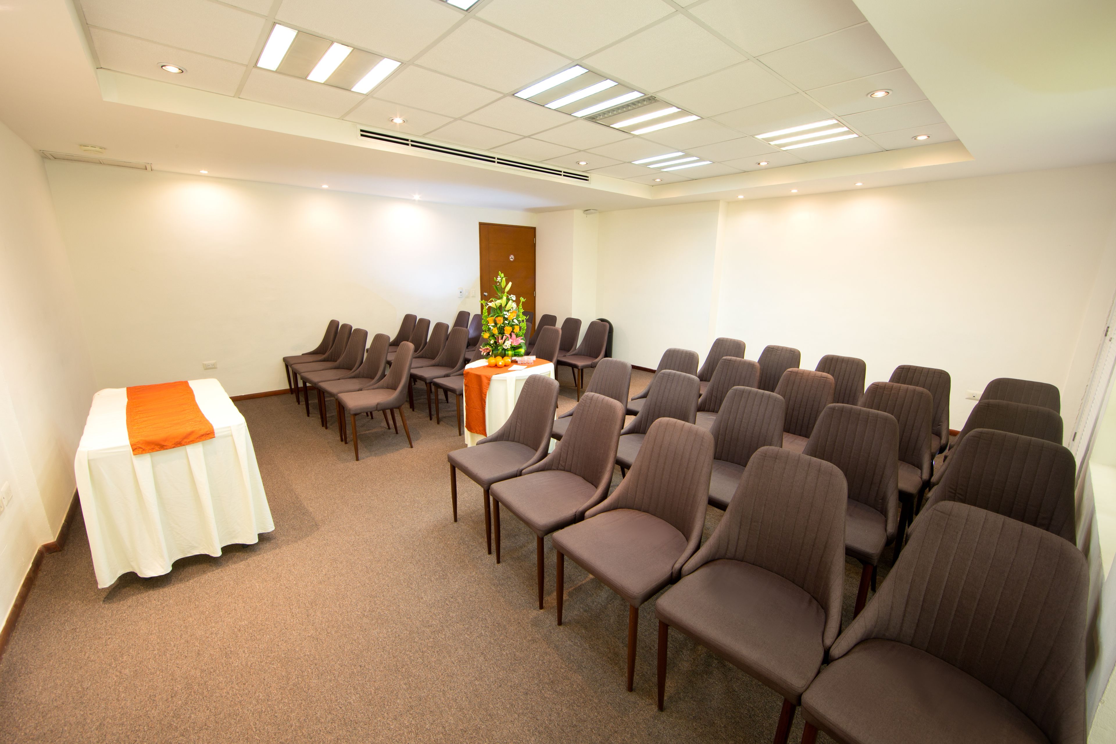 Ein Konferenzraum mit Reihen von Stühlen, einem Tisch mit weißem Tuch und einer orangefarbenen Tischläufer sowie einer Blumenkomposition.