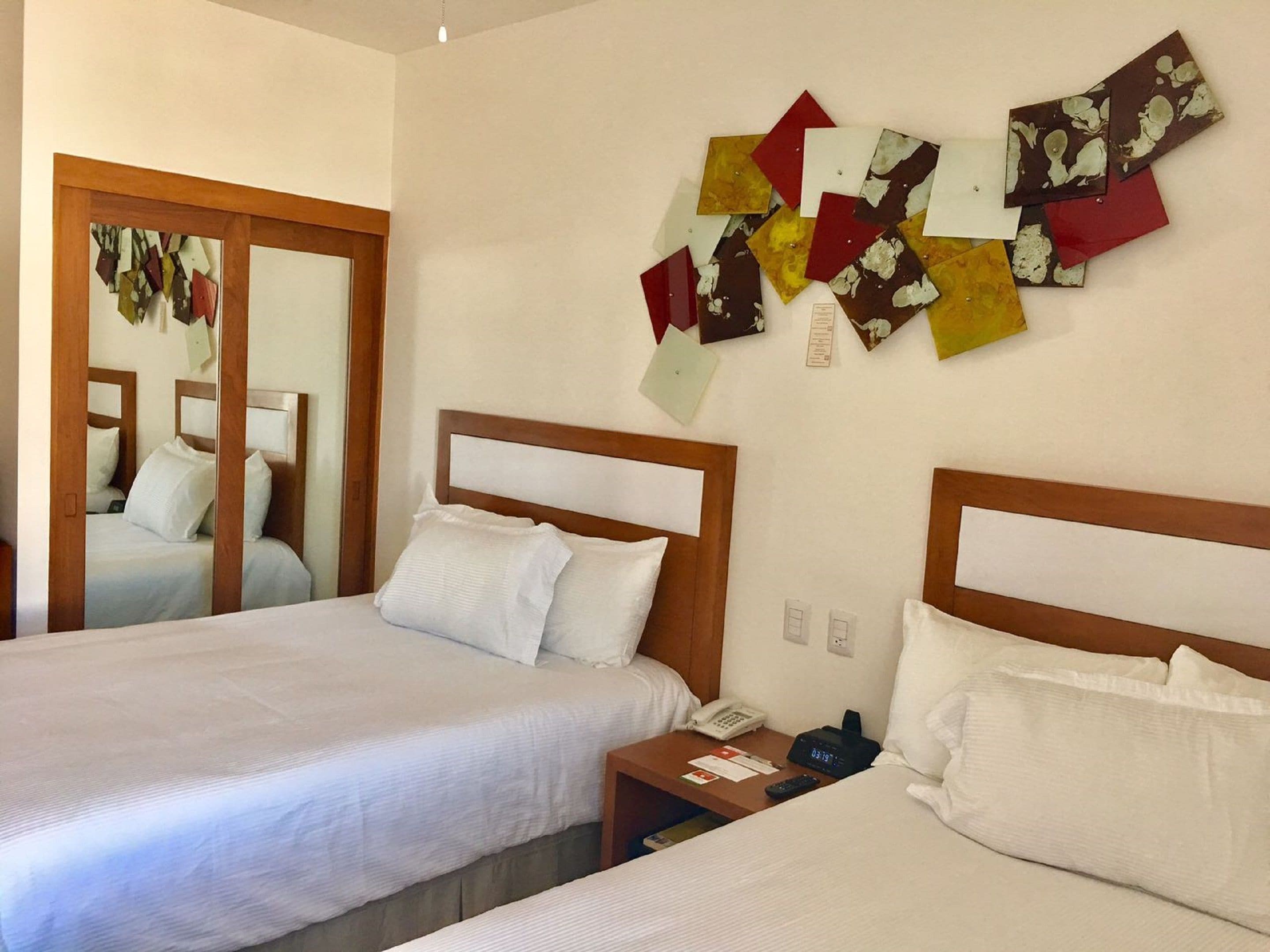 Una habitación de hotel con dos camas, un cabecero de madera, una mesita de noche con teléfono y reloj, y una pared decorada con arte colorido.