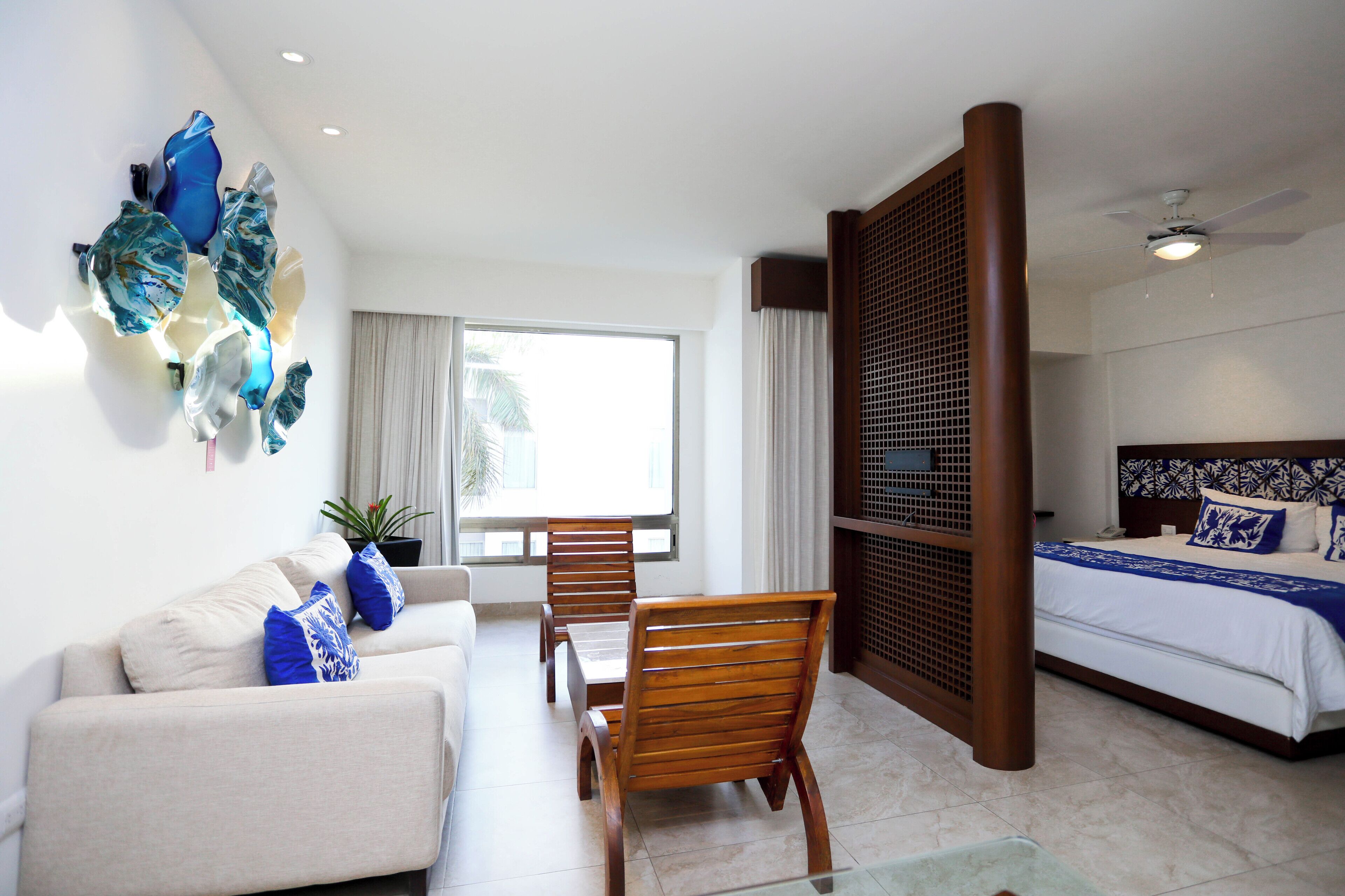 A hotel room with a bed, a sofa, a dining table, and chairs. There is a large window providing natural light and a view of palm trees.