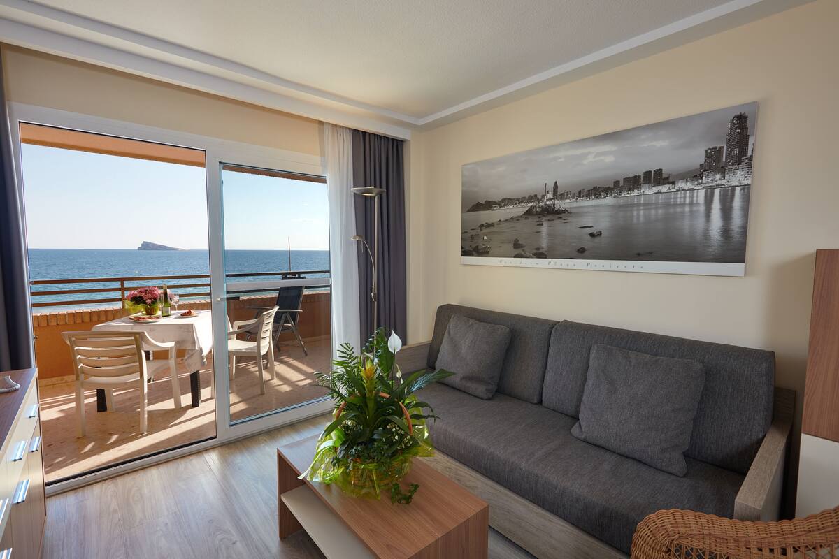 A modern living room with a grey sofa, a wooden coffee table, and a balcony with a table and chairs. A cityscape painting is on the wall, and a plant is on the coffee table.
