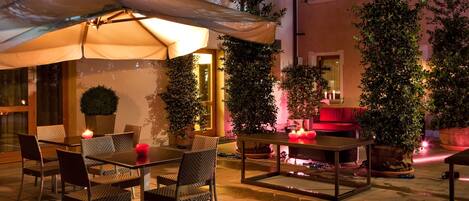 An outdoor patio with tables and chairs, surrounded by potted plants and a red-lit seating area.