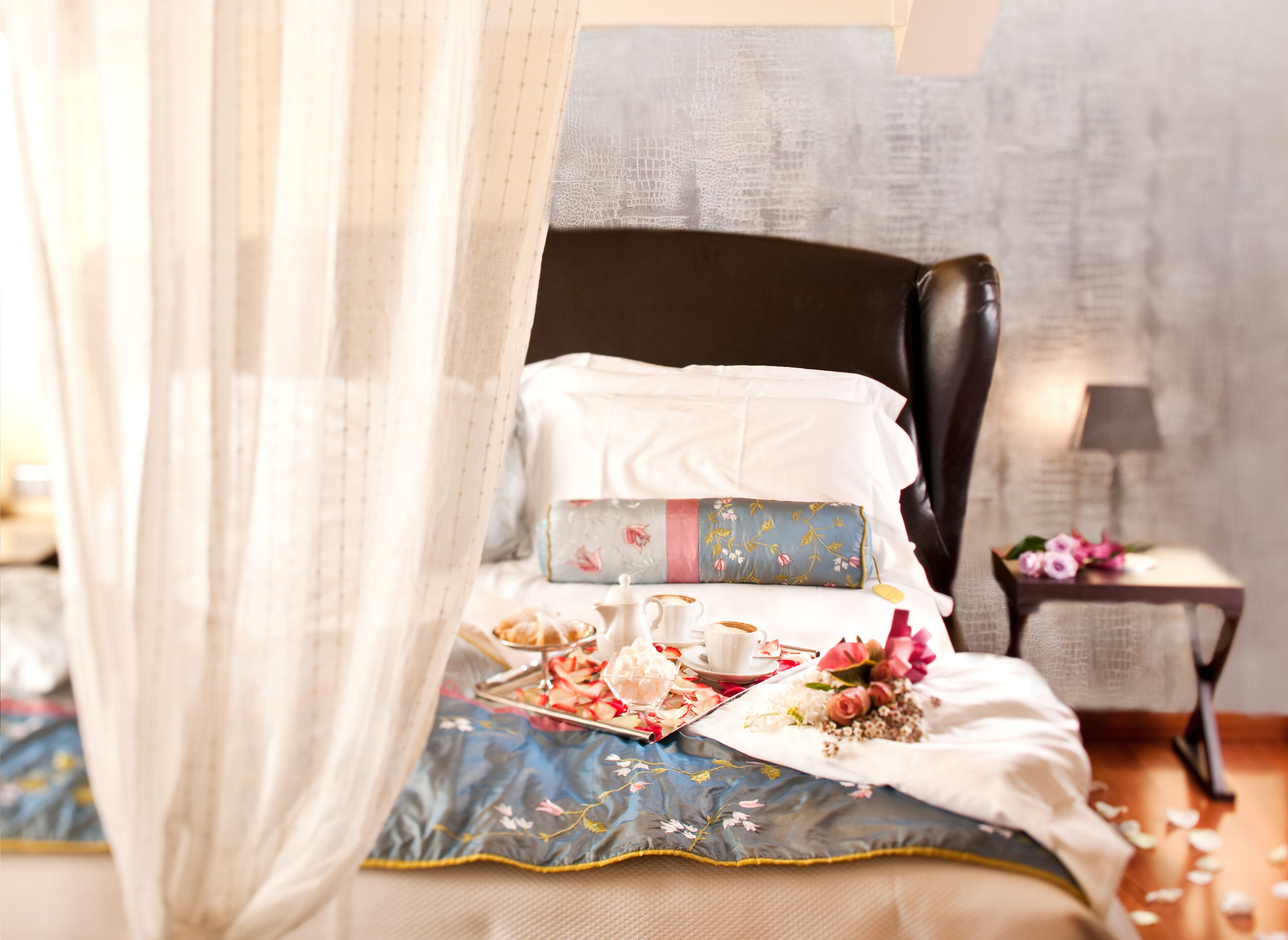 A neatly made bed with a canopy, floral bedding, and a bedside table with a lamp and flowers.