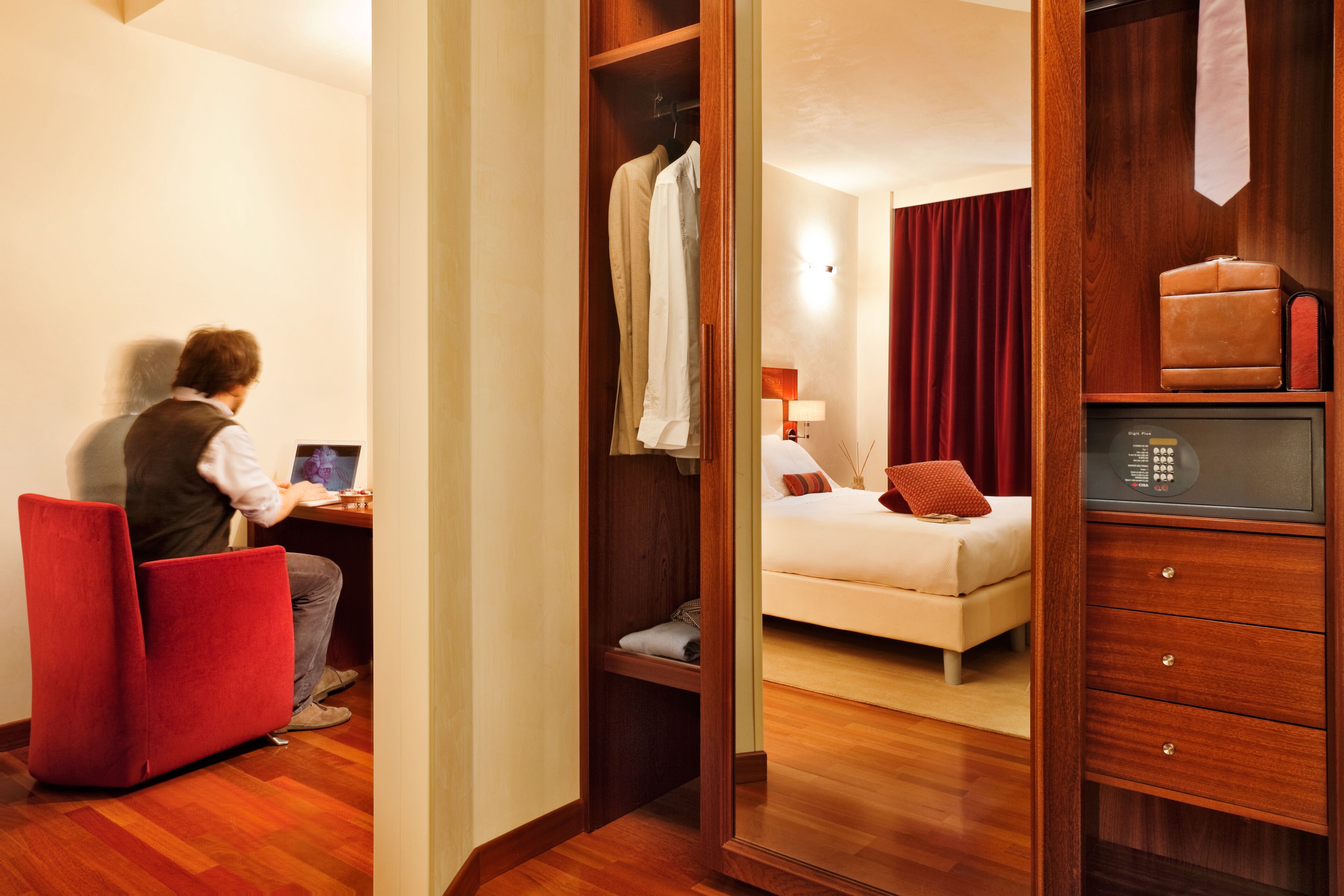 Una habitación de hotel con un escritorio de madera, una silla roja, una cama con cortina roja y un armario con una maleta.