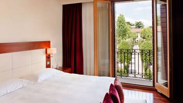 Habitación de hotel con una cama grande, una mesita de noche, una lámpara, una puerta corrediza de vidrio a un balcón y vistas al exterior.