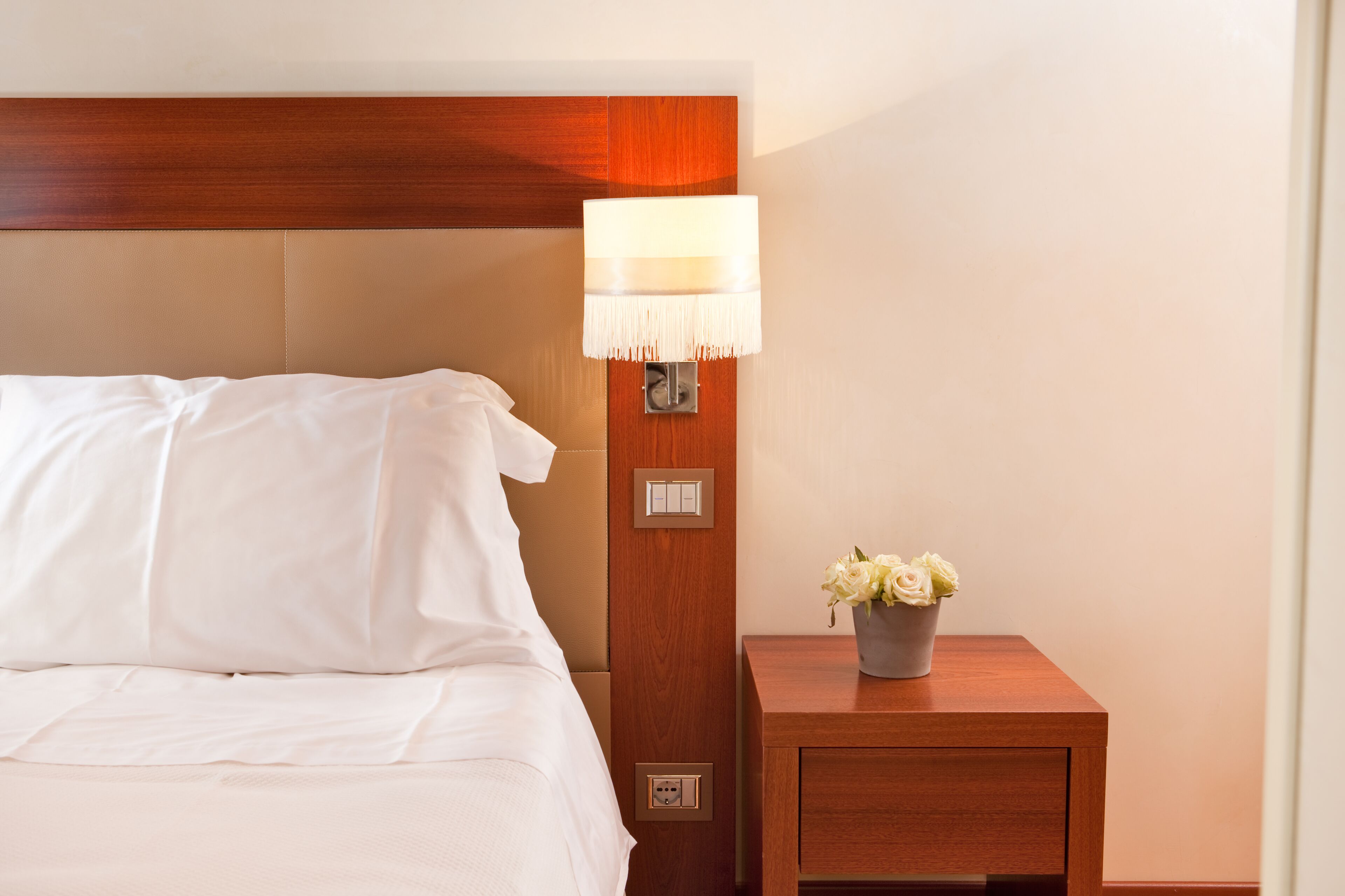 Ein ordentlich bezogenes Bett mit weißen Leinen, ein Nachttisch aus Holz mit einer Lampe und eine kleine Vase mit Blumen.